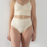 Model wearing High Meara organic cotton Brazilian brief, front view showing high-rise waist and leg-flattering cut. Cream