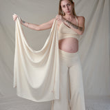 A woman modeling a matching cream loungewear set consisting of a cropped tank top, wide-leg pants, and a large draped fabric throw, posed against a neutral draped fabric backdrop.