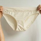 Front view of Tegan Full Bikini Brief in cream organic cotton, held delicately in the air by fingers. Shows soft fabric, full coverage cut, and ethical craftsmanship.